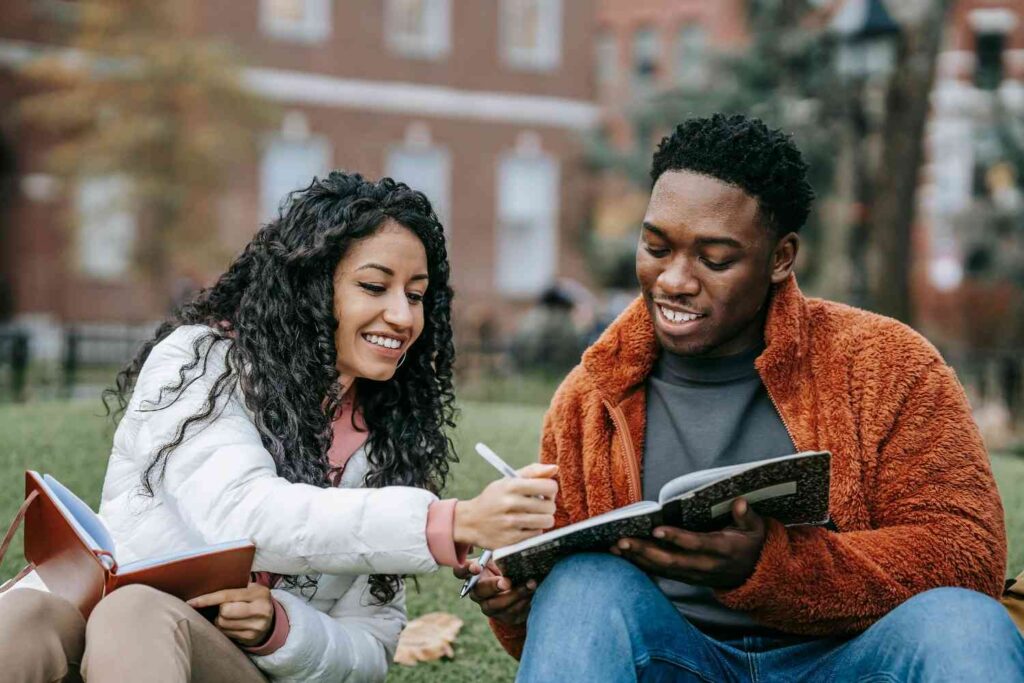 two students holding notebooks while sitting on grass
