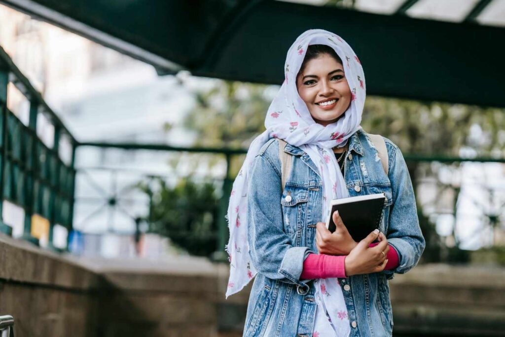 A smiling woman wearing a hijab carrying a notebook in her arms