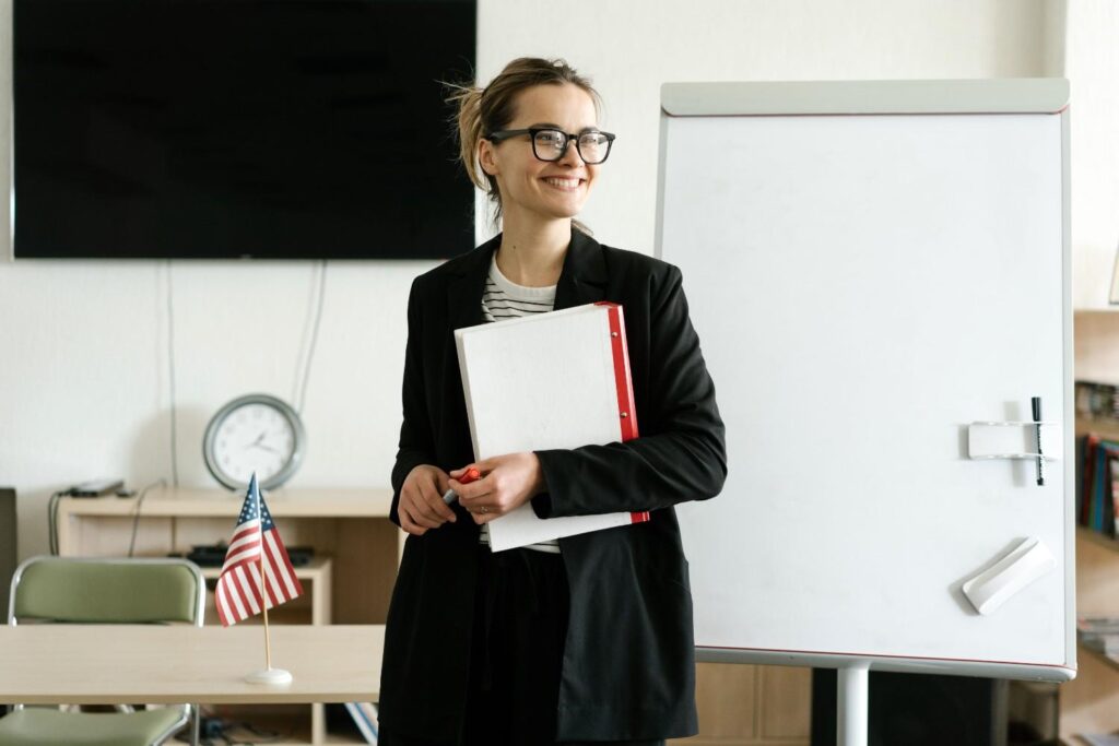 Smiling woman in black blazer holding notebooks next to a whiteboard with a US flag