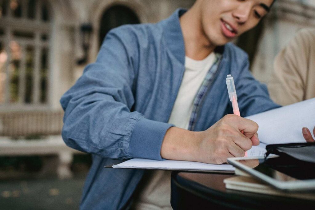 A young man concentrates on writing notes outdoors in an academic campus