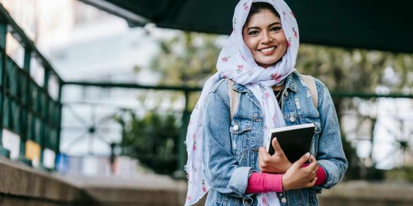 A smiling woman wearing a hijab carrying a notebook in her arms