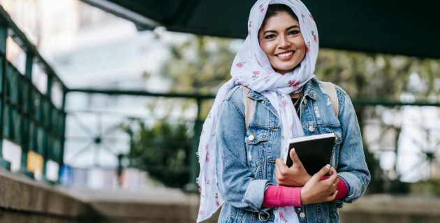 A smiling woman wearing a hijab carrying a notebook in her arms